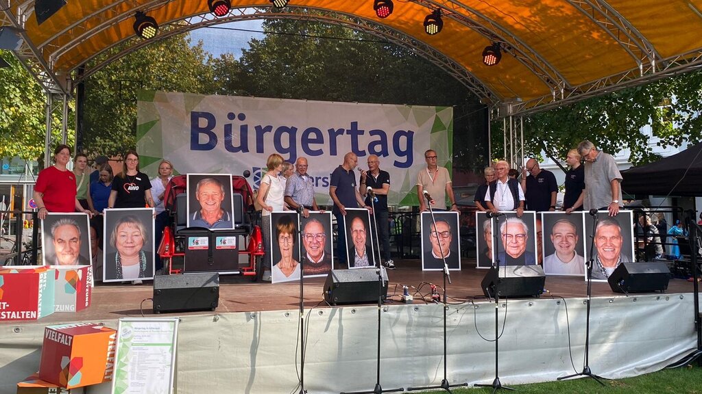 Mehrere Menschen stehen auf einer Bühne und halten große Portraitsfotos vor sich.