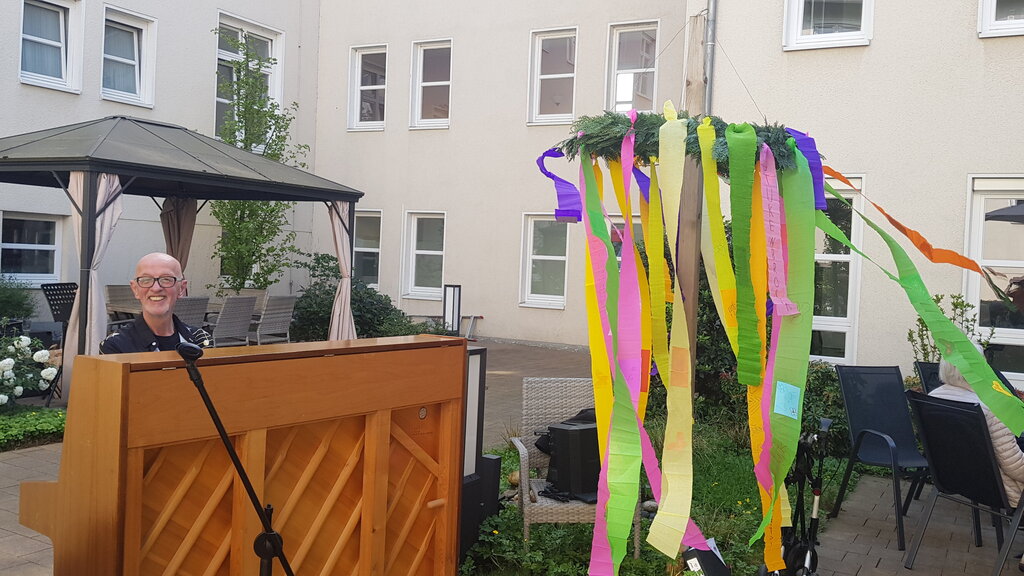 Mann am Klavier. Daneben ein bunter Maibaum.